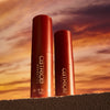 Zwei Catrice Cosmetics MIDNIGHT SUN Jelly Glow Sticks sind auf einer strukturierten Oberfläche platziert, vor dem Hintergrund eines Sonnenuntergangshimmels.