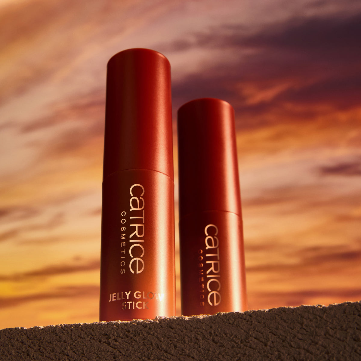 Zwei Catrice Cosmetics MIDNIGHT SUN Jelly Glow Sticks sind auf einer strukturierten Oberfläche platziert, vor dem Hintergrund eines Sonnenuntergangshimmels.