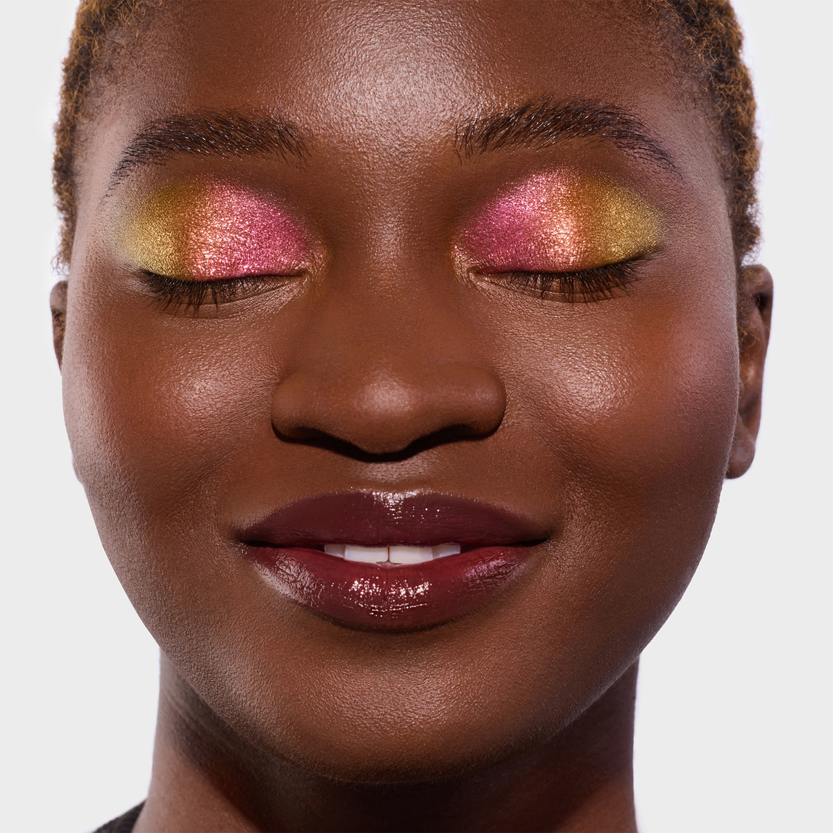 Eine Frau mit dunklem Hautton und kurzem Haar trägt rosa-orange und gelb schimmernden Twin Flames Lidschatten auf den Augen.