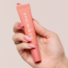 Eine Hand mit rosa Nägeln hält einen Juicy Lip Balm von Dominique Cosmetics vor einem hellen Hintergrund.