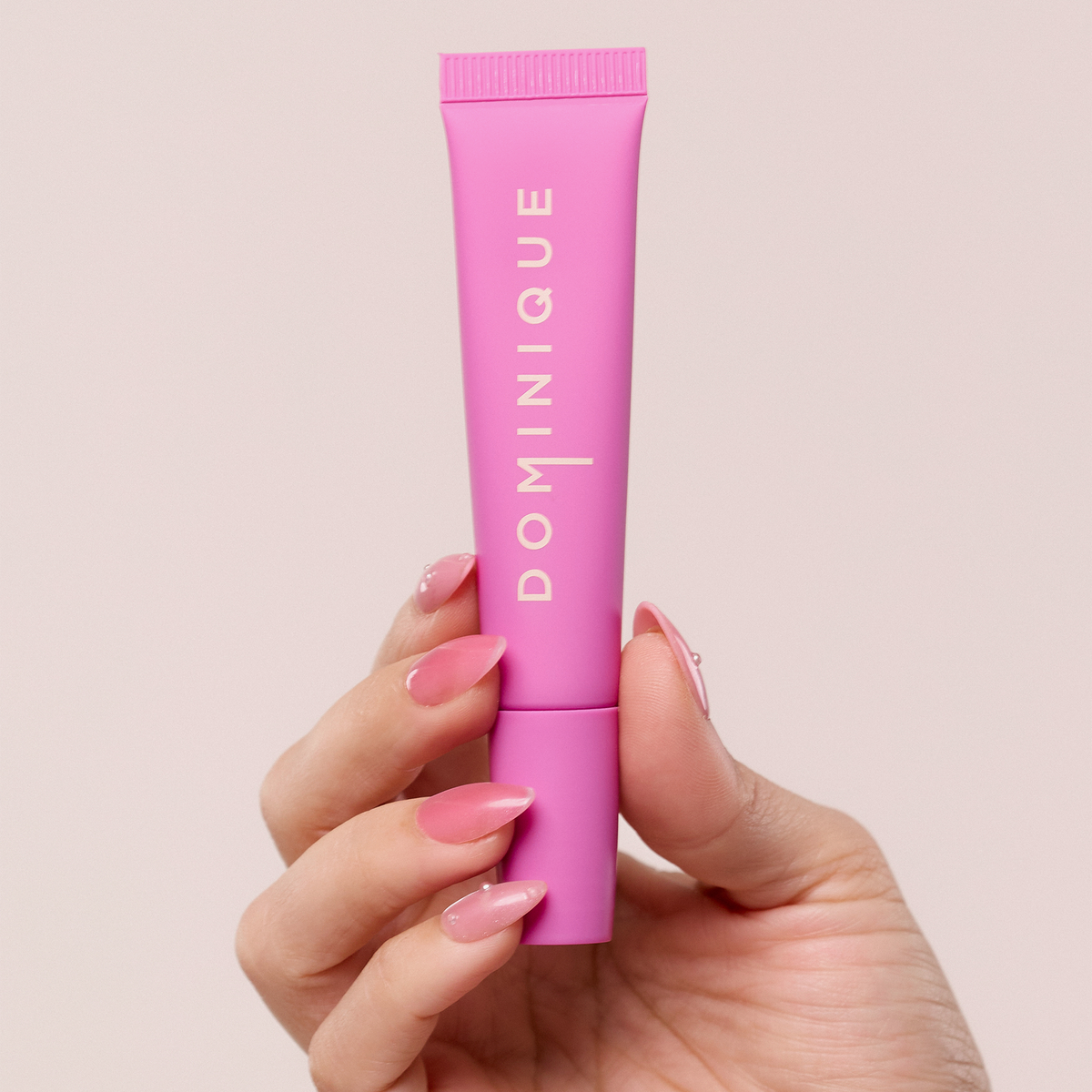 Eine Hand mit rosafarbenen Nägeln hält eine rosafarbene Tube Juicy Lip Balm von Dominique Cosmetics vor einem hellen Hintergrund.