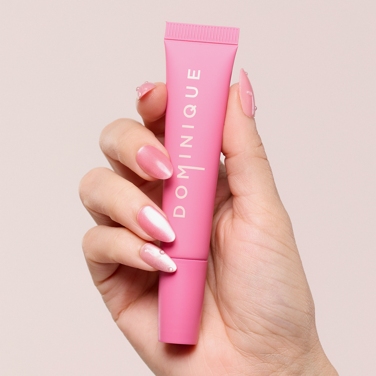 Eine Hand mit rosafarbenen manikürten Nägeln hält eine rosafarbene Dominique Cosmetics Juicy Lip Balm Tube vor einem hellen Hintergrund.