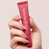 Eine Hand mit rosa Nägeln hält einen rosa Juicy Lip Balm von Dominique Cosmetics vor einem hellen Hintergrund.