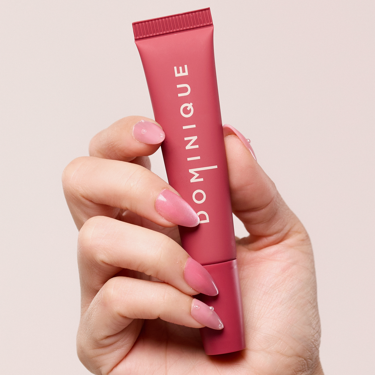 Eine Hand mit rosa Nägeln hält einen rosa Juicy Lip Balm von Dominique Cosmetics vor einem hellen Hintergrund.