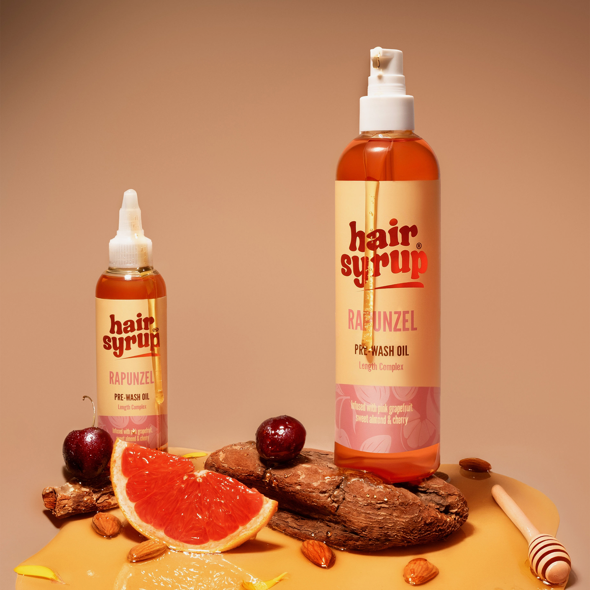 Zwei Flaschen Hair Syrup Rapunzel Pre-Wash Oil mit Grapefruit, Kirsche und Honig stehen auf einer braunen Fläche.