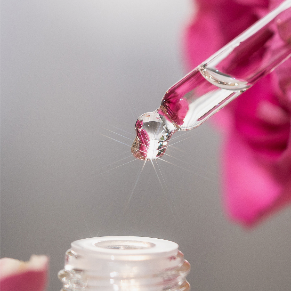 Ein Tropfer mit klarem Serum schwebt über einer Flasche Hair Syrup Rose Bloom, mit einer verschwommenen rosa Blume im Hintergrund.