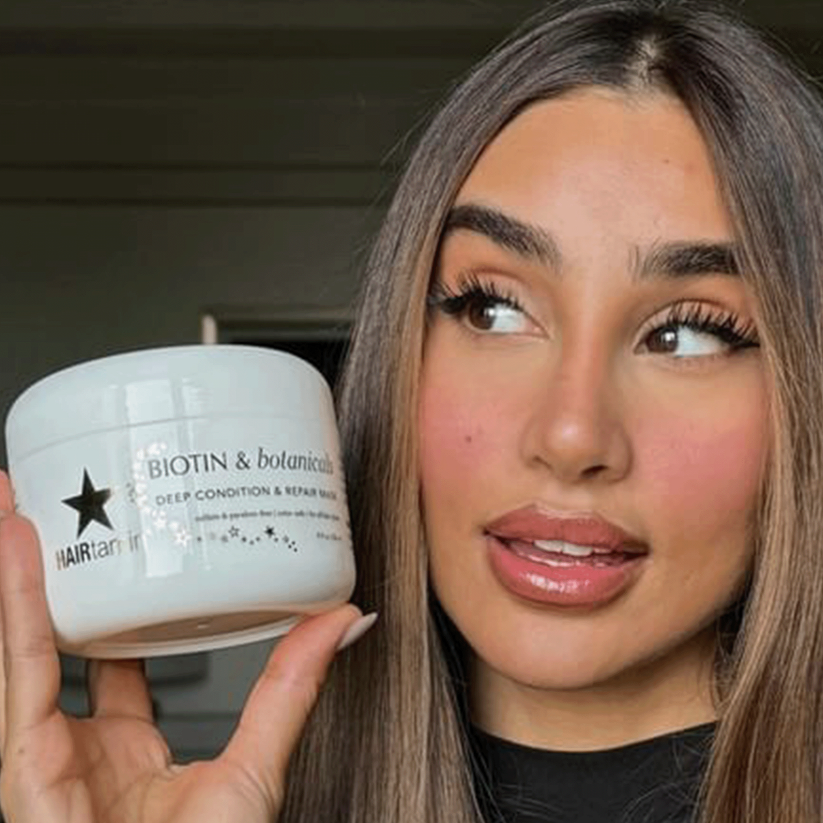 Frau hält einen Tiegel der Hairtamin Biotin & Botanicals Deep Condition & Repair Haarmaske neben ihr Gesicht, natürliches Licht, gepflegtes Haar.