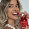 Eine lächelnde Frau, die eine rote Flasche mit der Aufschrift „HAIRtamin Gummy Stars™ 3-Monats-Paket“ in der Hand hält und mit der anderen Hand ein Gummibonbon isst.