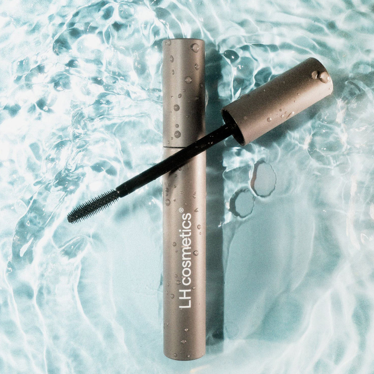 Graue Mascara mit geöffnetem Deckel und sichtbarer Bürste liegt auf einer Wasseroberfläche mit Lichtreflexen; Wassertropfen bedecken die Verpackung.