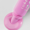 Lumene Lip Glow Oil Balm SPF30 in 2 Marshmallow wird aus der Tube gedrückt und bildet eine glänzende rosa Pfütze auf einer weißen Oberfläche.