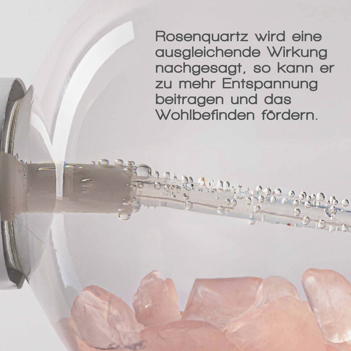 Nahaufnahme einer Sprühflasche mit Rosenquarzkristallen im Inneren und Luftblasen im Schlauch. Text: „Rosenquarz wird eine ausgleichende Wirkung nachgesagt …“