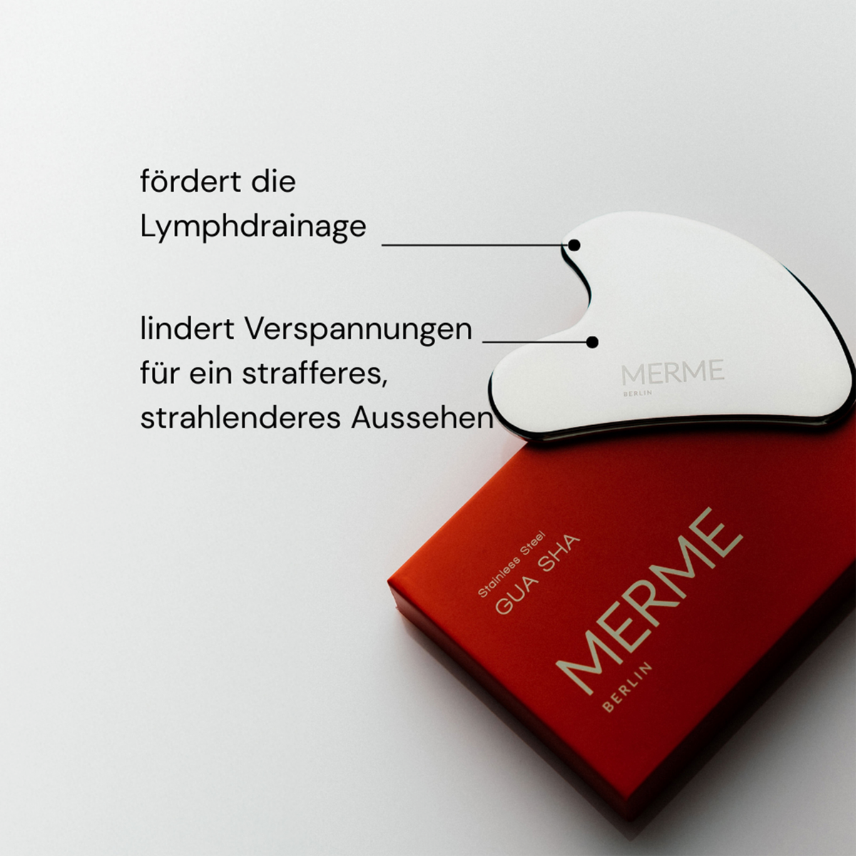 Gua Sha aus Edelstahl von MERME mit beschrifteten Vorteilen, auf einer roten Schachtel, vor einem hellen Hintergrund.