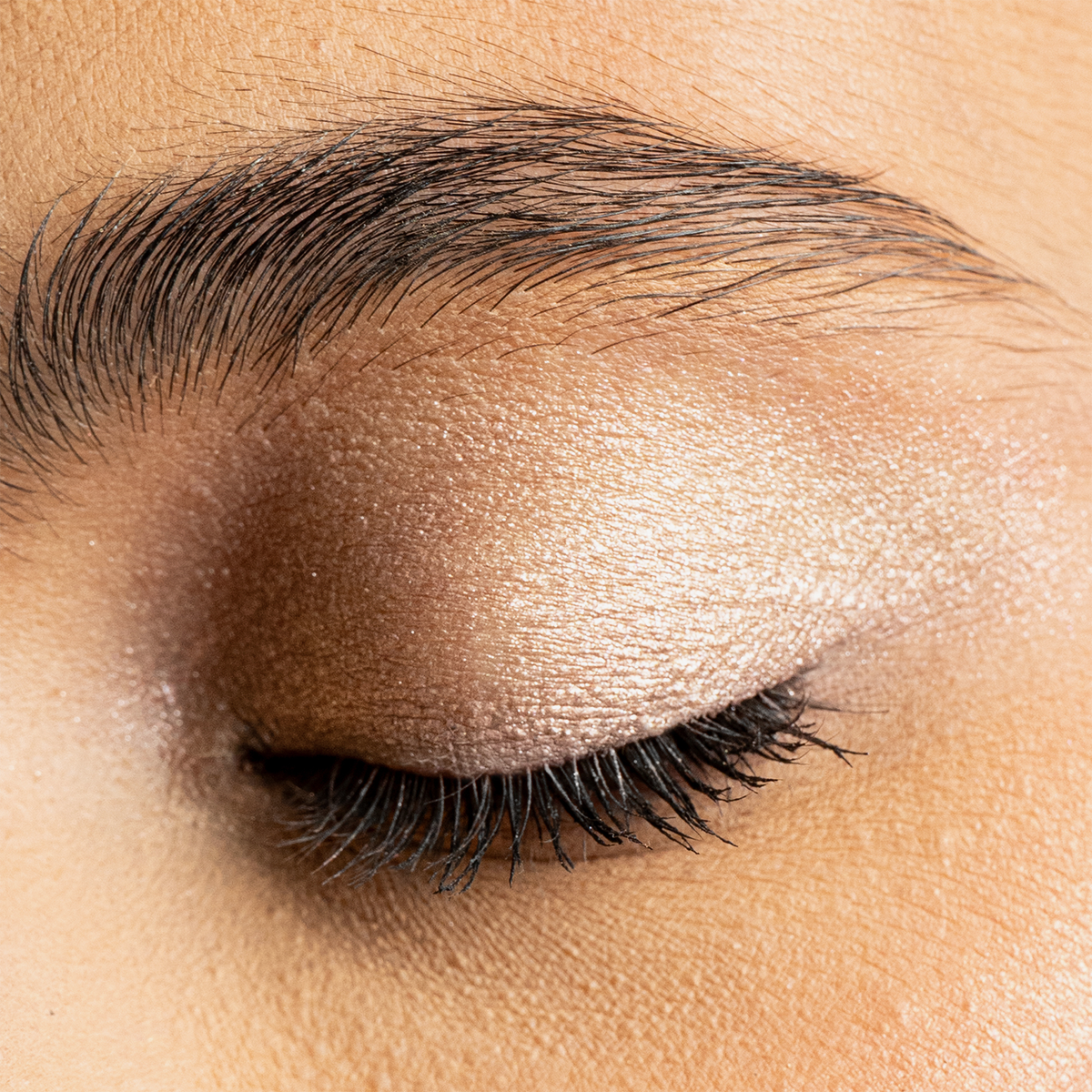 Nahaufnahme eines geschlossenen Auges mit schimmerndem, champagnerfarbenem Lidschatten und betonten schwarzen Wimpern.