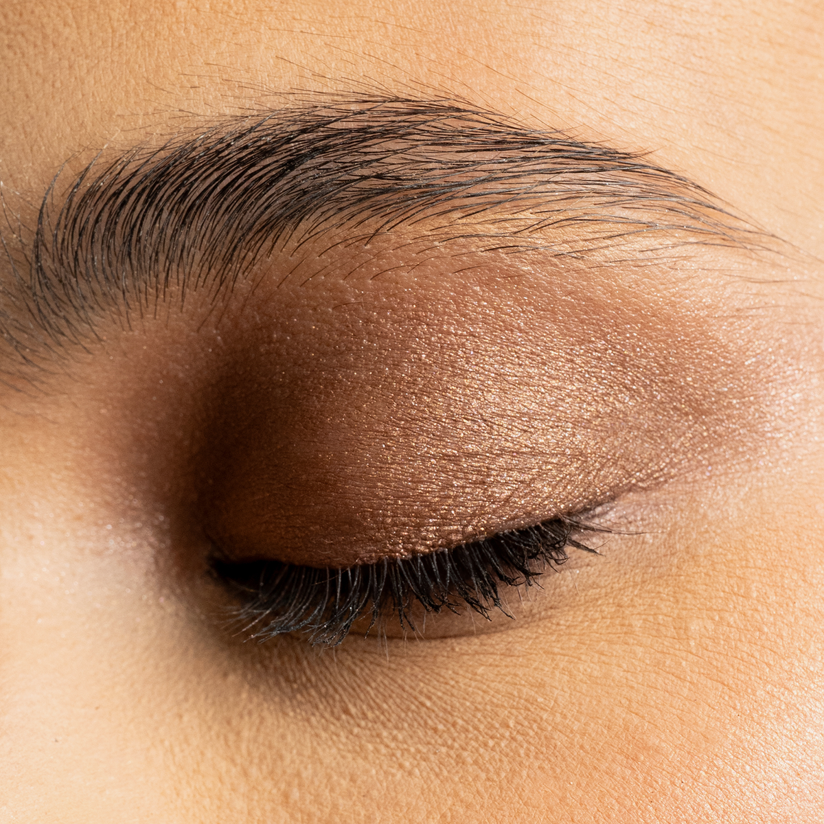 Nahaufnahme eines geschlossenen Auges mit warmem, bronzefarbenem Lidschatten in satiniertem Finish und betonten schwarzen Wimpern.