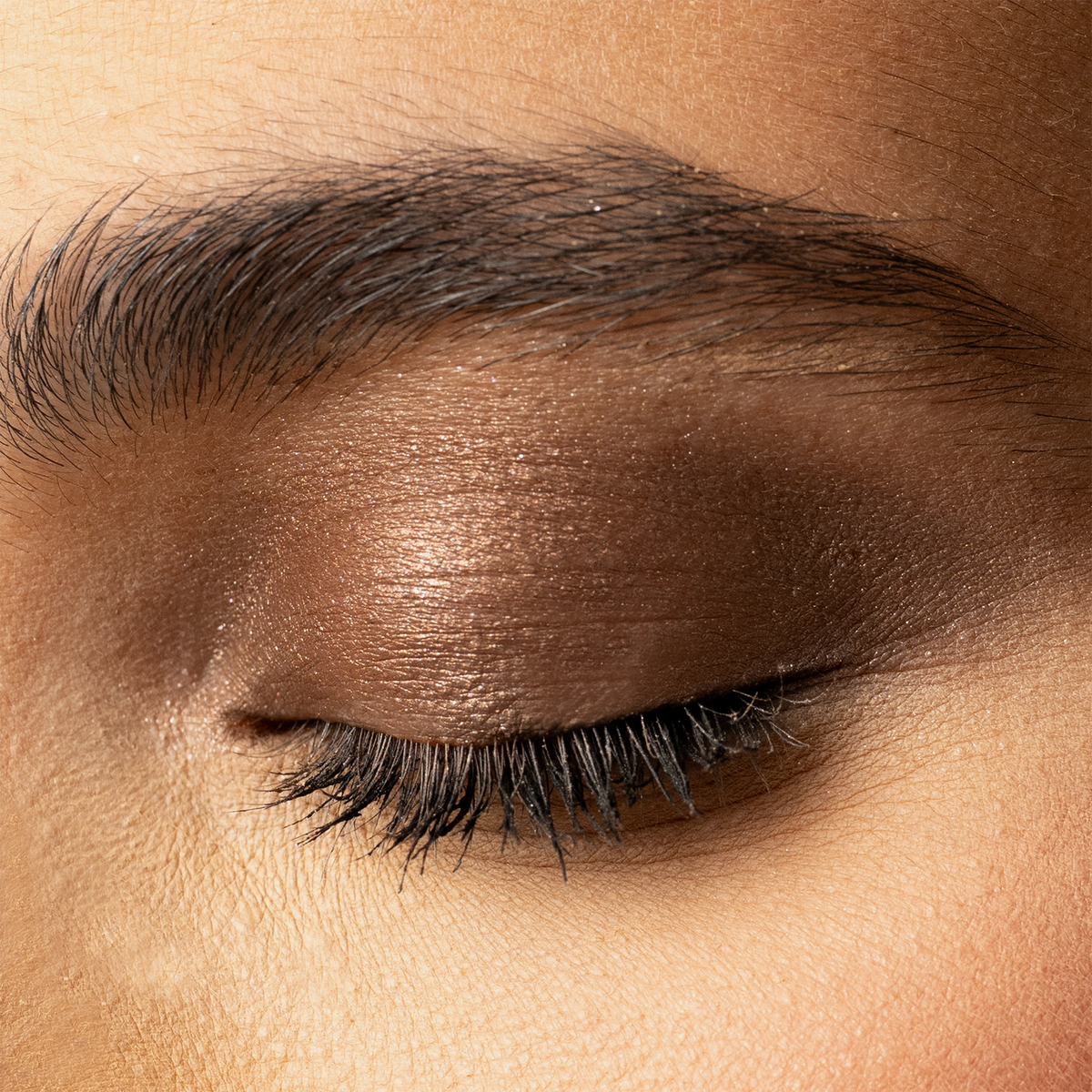Nahaufnahme eines geschlossenen Auges mit schimmerndem bronzefarbenem Lidschatten in warmem Ton und betonten schwarzen Wimpern.