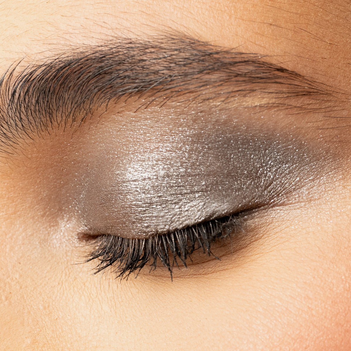 Nahaufnahme eines geschlossenen Auges mit metallisch-grauem, rauchigem Lidschatten in kühlem Ton und definierten schwarzen Wimpern.