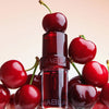 NABLA Cosmetics Lip Candy Oil Black Cherry, umgeben von glänzenden Kirschen vor weichem Farbverlauf-Hintergrund.