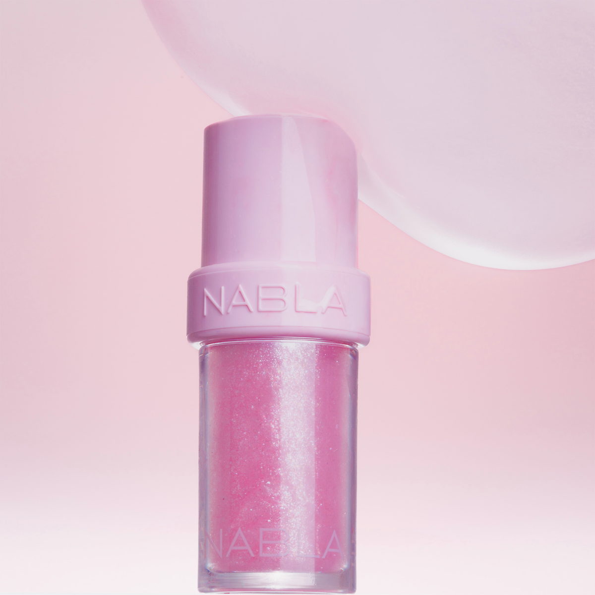 Ein „Lip Candy Oil“ von NABLA Cosmetics steht aufrecht auf einem zartrosa Hintergrund, über dem sich eine flauschige Form abzeichnet.