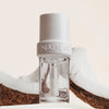 Transparentes Fläschchen NABLA Cosmetics Lip Candy Oil Salted Coconut, umgeben von Kokosnussschalen vor hellem Hintergrund.