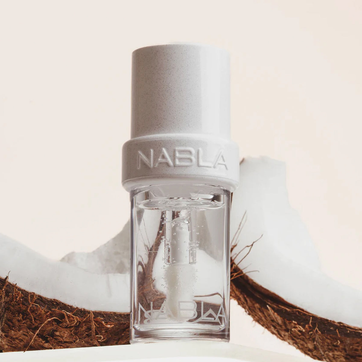 Transparentes Fläschchen NABLA Cosmetics Lip Candy Oil Salted Coconut, umgeben von Kokosnussschalen vor hellem Hintergrund.