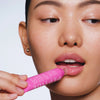 Eine Person trägt Paula's Choice Pro-Collagen Peptide Gloss Balm auf die Lippen auf und präsentiert glatte, strahlende Haut vor einem neutralen Hintergrund.
