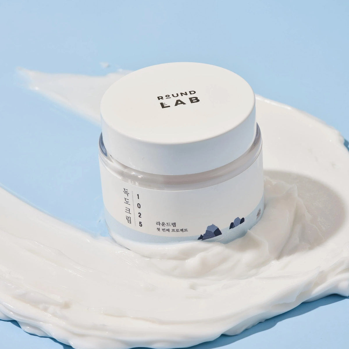 Runder Tiegel der Round Lab 1025 Dokdo Cream auf hellblauem Hintergrund, umgeben von aufgetragener weißer Creme mit weicher, geschmeidiger Textur.