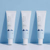 Drei Tuben der Round Lab 1025 Dokdo Eye Cream stehen aufrecht nebeneinander vor hellblauem Hintergrund.