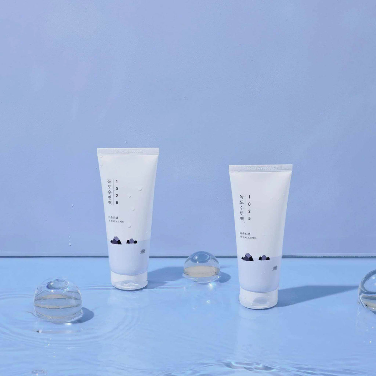 Zwei Tuben des Round Lab 1025 Dokdo Sleeping Pack stehen in Wasser, umgeben von klaren Gel-Kugeln, inszeniert vor blauem Hintergrund.