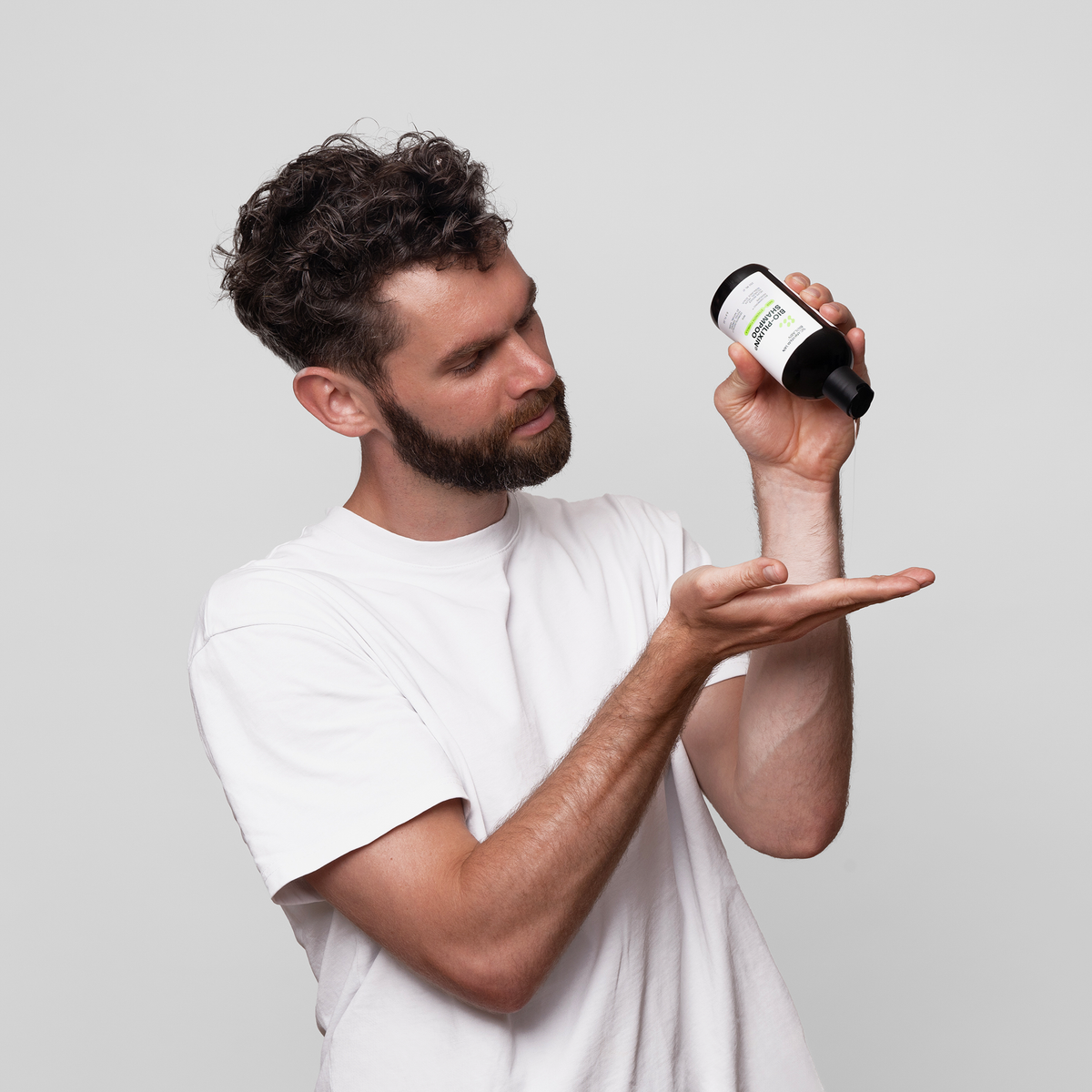 Ein Mann gießt Bio-Pilixin® Shampoo+ Men von Scandinavian Biolabs aus einer dunklen Flasche in seine Hand, vor einem schlichten Hintergrund.