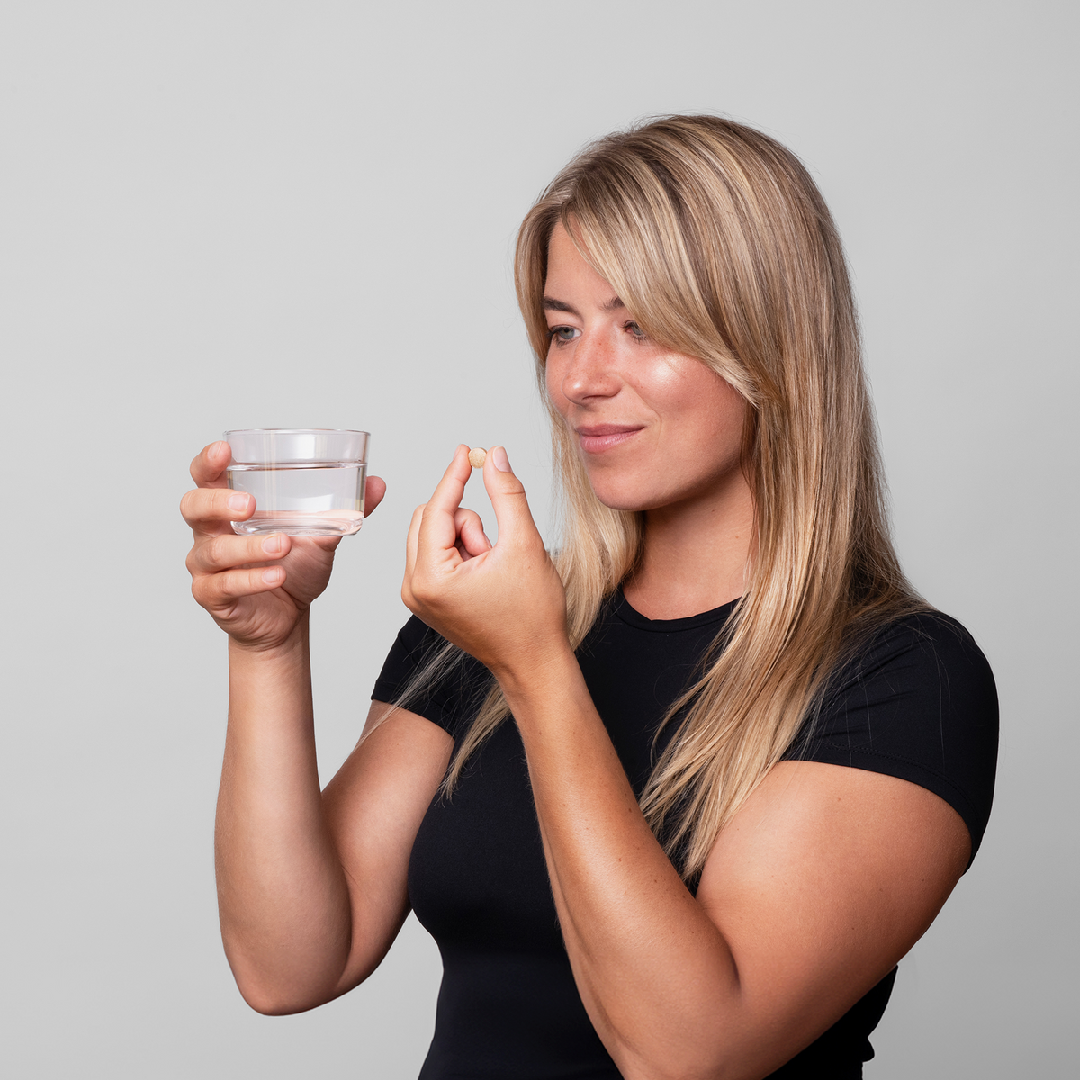 Eine Frau hält ein Glas Wasser und eine Hair Nutrient Tablette von Scandinavian Biolabs in der Hand und ist im Begriff, das Präparat einzunehmen.