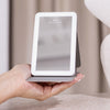 Eine Hand hält den STYLPRO LED Mini Mirror, einen kompakten beleuchteten Spiegel mit klappbarem Ständer, auf einer beigen Oberfläche.