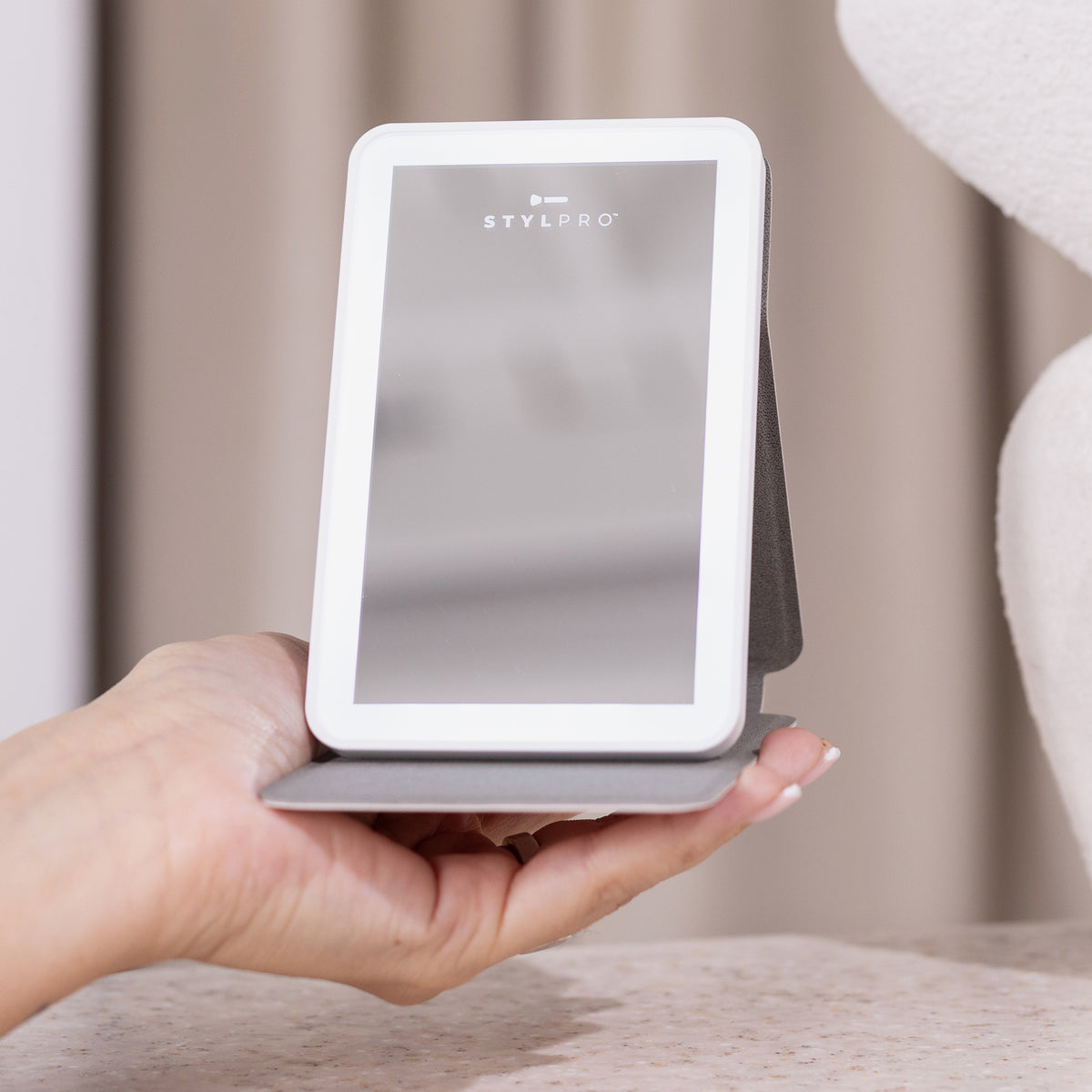 Eine Hand hält den STYLPRO LED Mini Mirror, einen kompakten beleuchteten Spiegel mit klappbarem Ständer, auf einer beigen Oberfläche.