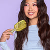 Lächelnde Frau mit langem gewelltem Haar hält eine gelbe UNbrush Detangling Hair Brush vor einem lila Hintergrund.