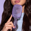 Eine Frau mit gewelltem braunem Haar hält die UNbrush Detangling Hair Brush in Lila vor einem lavendelfarbenen Hintergrund an ihr Kinn.