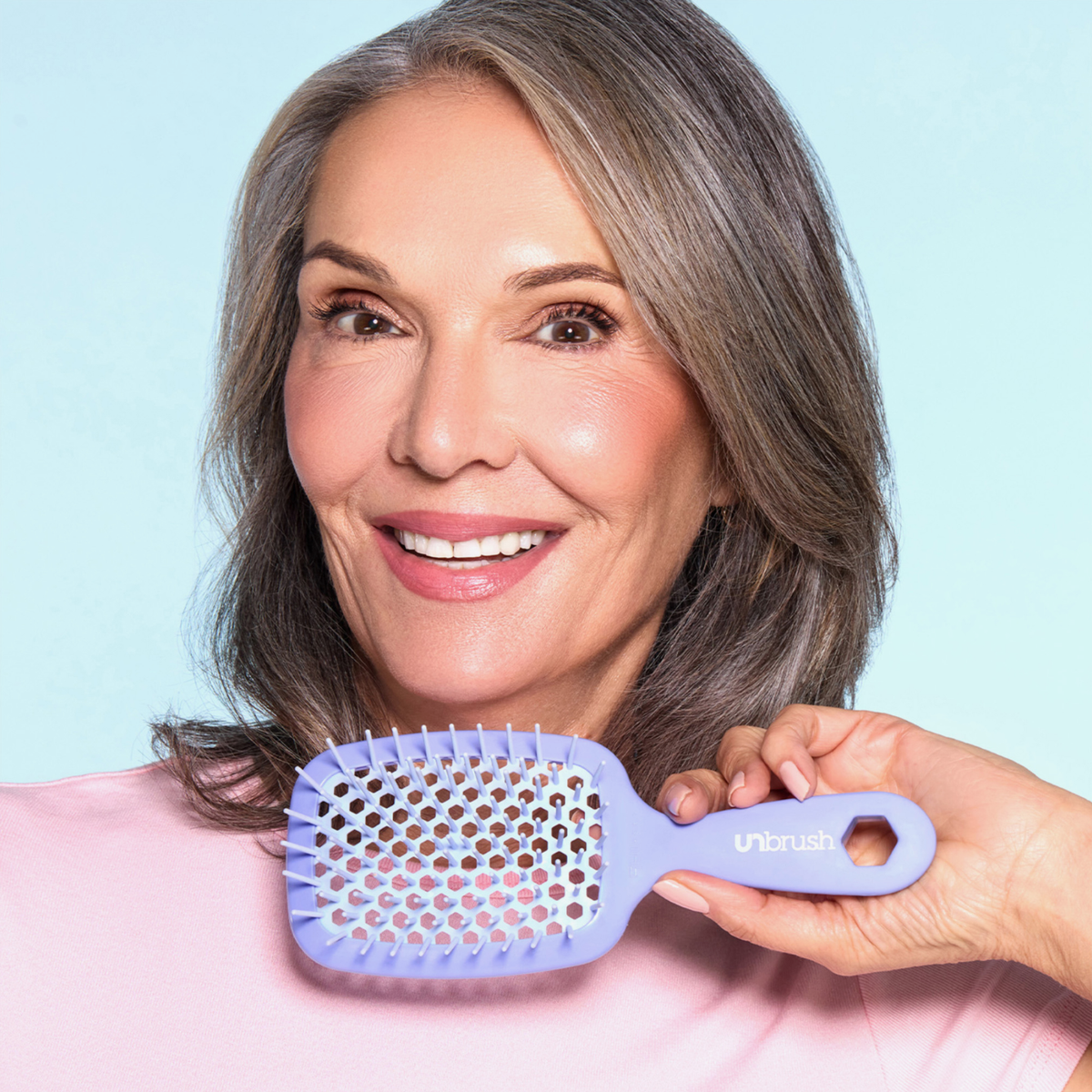 Eine lächelnde grauhaarige Frau hält die UNbrush Detangling Hair Brush vor einem hellblauen Hintergrund.