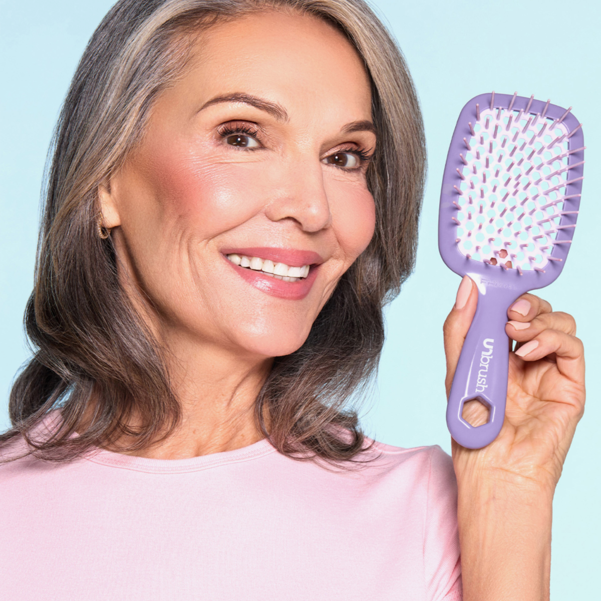 Lächelnde Frau mit grauem Haar hält eine UNbrush Detangling Hair Brush vor einem hellblauen Hintergrund.