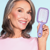 Lächelnde Frau mit grauem Haar hält die UNbrush Detangling Hair Brush vor einem hellblauen Hintergrund.