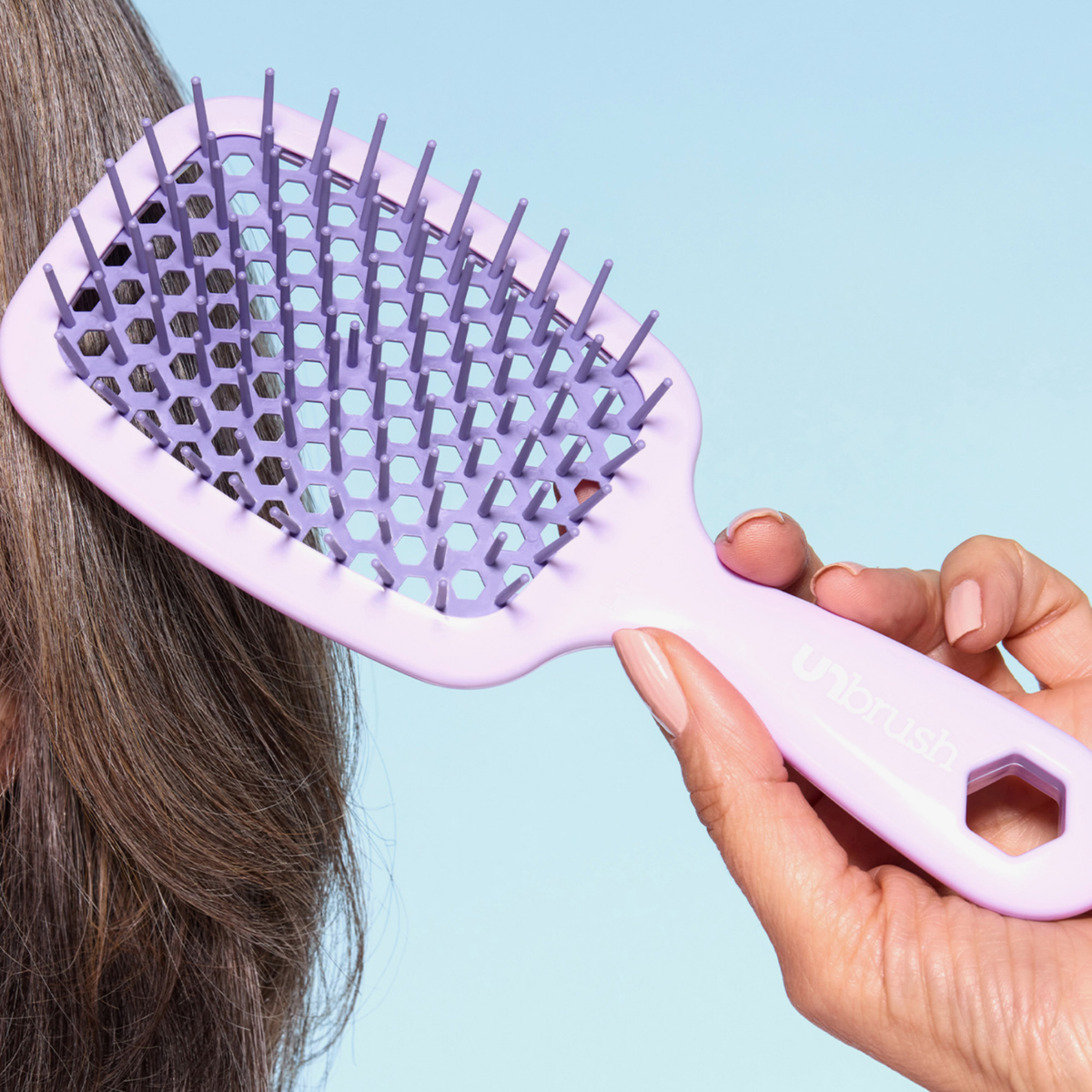 Eine Hand bürstet graues Haar mit der UNbrush Detangling Hair Brush Mini (hellviolett) vor einem blauen Hintergrund.