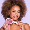 Lächelnde Frau mit lockigem Haar hält die UNbrush Detangling Hair Brush Mini, trägt helles Make-up und ein rosa Oberteil.