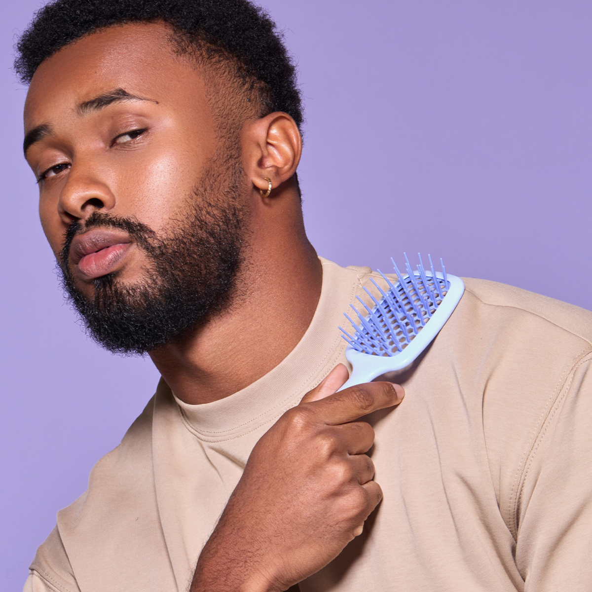 Ein Mann benutzt die UNbrush Detangling Hair Brush Mini im Nacken. Er trägt Beige und hat einen violetten Hintergrund vor sich.