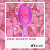 Eine rosafarbene Detangling-Haarbürste Mini von UNbrush ruht auf Rosenquarzsteinen, auf denen unten "ROSE QUARTZ MINI" und das UNbrush-Logo zu sehen sind.