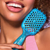 Eine lächelnde Frau in einem rosafarbenen Oberteil hält die UNbrush Detangling Hair Brush Mini, eine blaue Bürste, in der Nähe ihrer lockigen Haare.