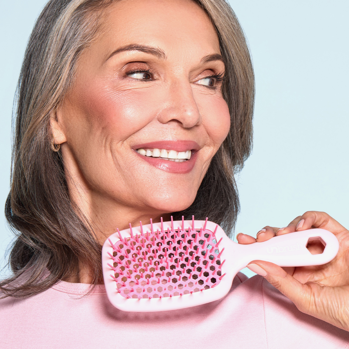 Lächelnde ältere Frau mit grauem Haar hält die UNbrush Detangling Hair Brush in der Nähe ihres Gesichts vor einem hellblauen Hintergrund.