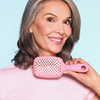 Eine lächelnde grauhaarige Frau hält eine rosa UNbrush Detangling Hair Brush in der Nähe ihres Gesichts vor einem hellblauen Hintergrund.