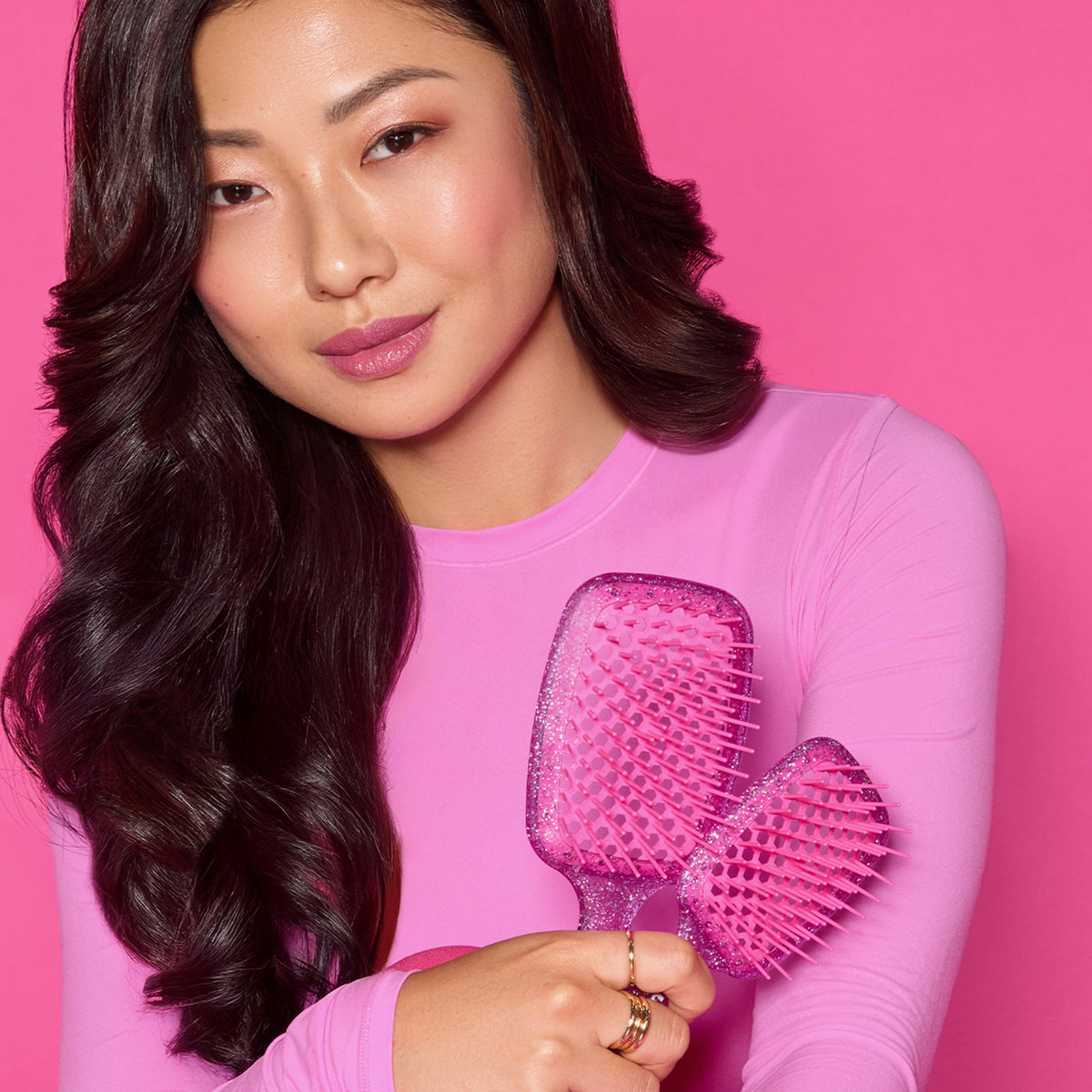 Eine Frau mit langen dunklen Haaren posiert vor einem hellrosa Hintergrund und hält zwei rosa glitzernde UNbrush Detangling Hair Brushes.