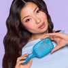 Eine Frau mit langem gewelltem Haar hält die UNbrush Detangling Hair Brush in Blau und posiert vor einem hellvioletten Hintergrund.