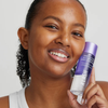 Eine lächelnde Person hält vor einem schlichten Hintergrund eine lila-weiße Flasche „XLASH Caring Eyes Waterproof Make-Up Remover“ in Größe XL nah an ihr Gesicht.