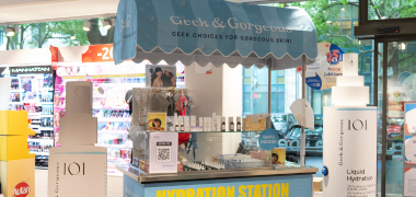 Geek & Gorgeous-Hautpflegestand mit blauer Markise, ausgestellten Produkten und dem Schild "Hydration Station" in einem hellen Geschäft.