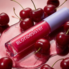 Eine Tube BLONDESISTER 2 IN 1 SWEET GLOSS TINTED LIP OIL steht zwischen frischen Kirschen auf einer glänzenden rosa Fläche.
