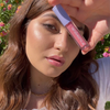 Eine Frau im Freien, die das „BLONDESISTER 2 IN 1 SWEET GLOSS TINTED LIP OIL“ nahe an ihr Gesicht hält, mit glänzenden Lippen und strahlender, sonnenbeschienener Haut.